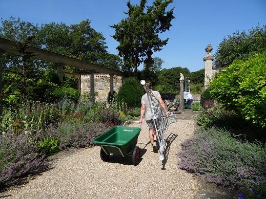Person carrying a ladder and pulling a wheelbarrow through a garden path